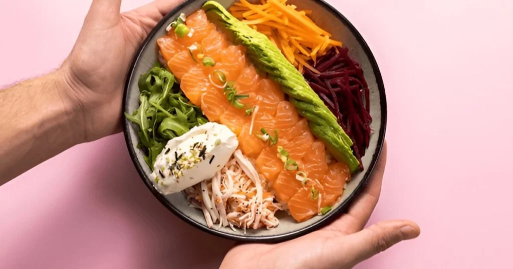 A vibrant salmon bowl featuring grilled salmon, steamed rice, colorful vegetables, and a drizzle of sesame sauce, garnished with sesame seeds and lime wedges.