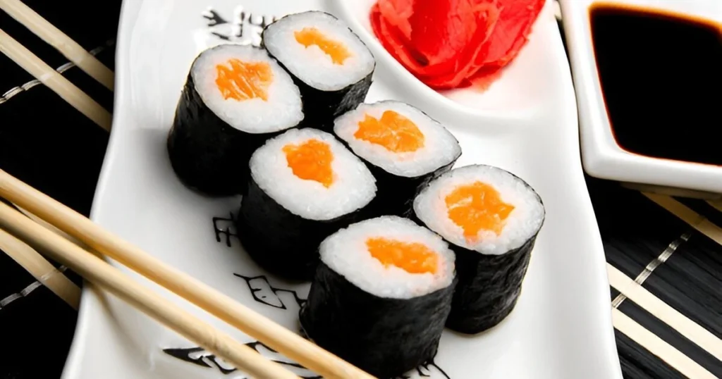 A close-up view of a baked sushi dish featuring tender salmon chunks, creamy layers of mayonnaise, and golden crispy edges, garnished with green onions and sesame seeds, served alongside nori sheets.