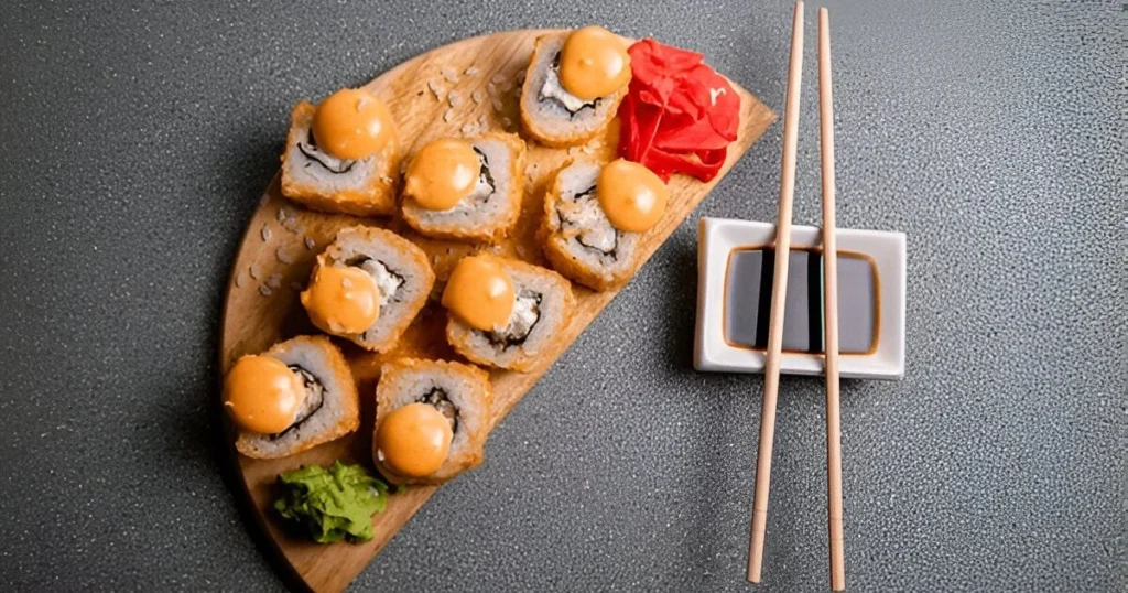 A close-up view of a baked sushi dish featuring tender salmon chunks, creamy layers of mayonnaise, and golden crispy edges, garnished with green onions and sesame seeds, served alongside nori sheets.